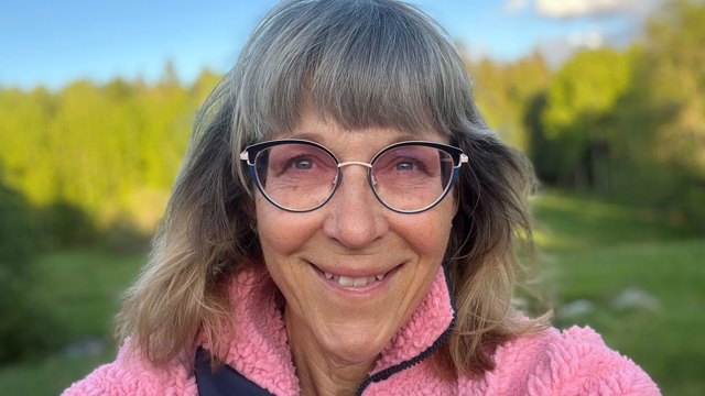 Leende kvinna tittar rakt in i kameran. Klädd i rosa tjocktröja. I bakgrunden syns natur och skogsmiljö.