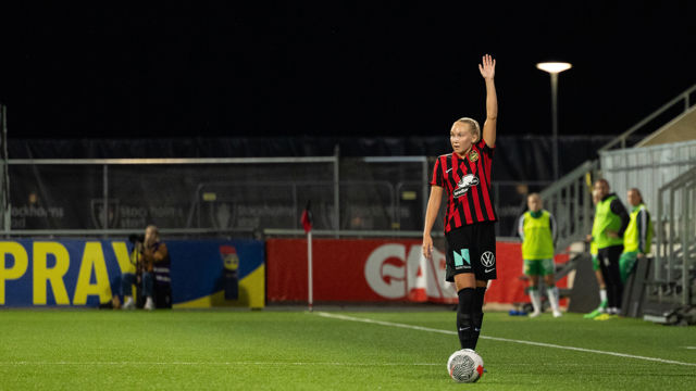 Kvinna på fotbollsplan, iklädd svart-röda kläder och stäcker upp ena armen.