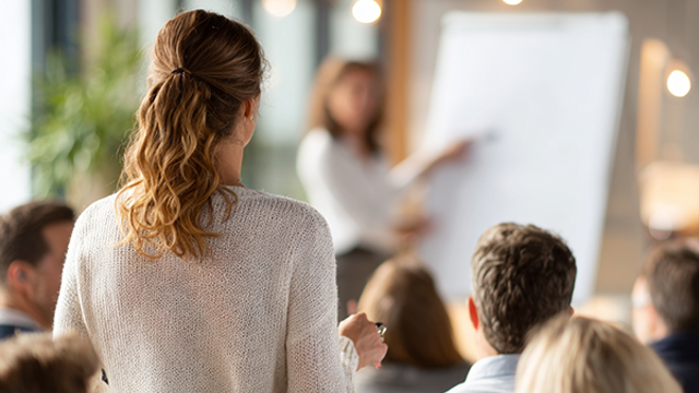 En person ställer en fråga under en presentation, där en föreläsare pekar på en whiteboard framför en grupp åhörare.